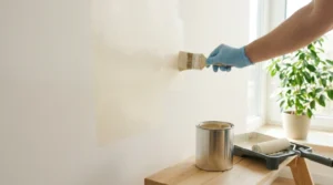 Person applying white paint on wall with brush beside open quart can and roller tray